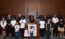 Children and Police Commanders proudly stand at a podium in front of a dias and flags.