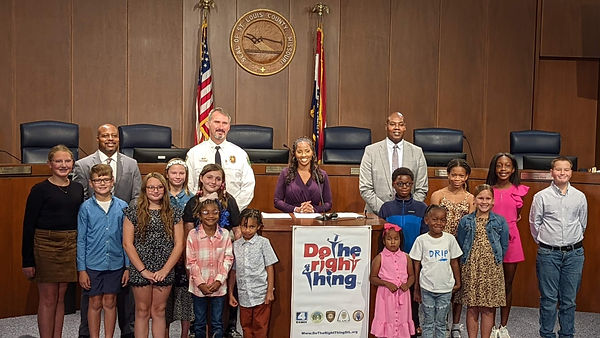 Fourteen amazing kiddos who are winners smile together in front of the St. Louis County Council Dias, flanked by police commanders, and KMOV Anchor Taylor Holt standing proudly in the middle behind the podium with the Do The Right Thing banner displayed on it.