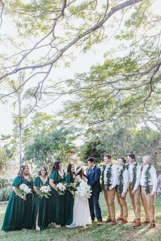 Bride and groom standing with their wedding party under tropical trees at Palikū Gardens, Kualoa Ranch in Oʻahu, laughing and holding floral leis and white bouquets.
