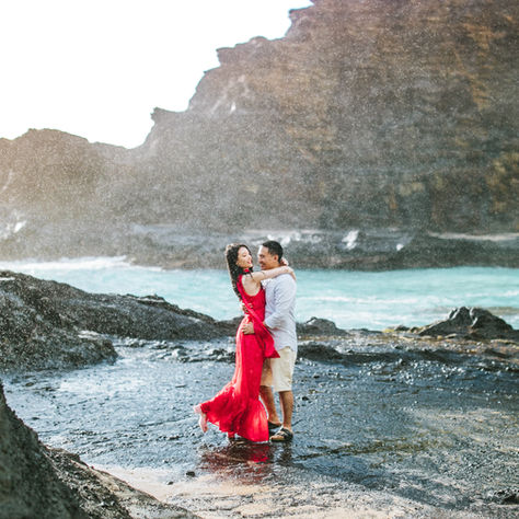 Couple during Hawaii photo shoot embracing on black lava rock with ocean waves and golden light, wearing a bold red dress that flows in the breeze