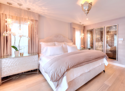 The luxurious Los Angeles  Primary Bedroom in a hotel style is a mushroom pink color, the walls, drapery and suede upholstered headboard all the same hue of color. Three antique mercury wall mirrors subtly reflect the courtyard into the moroccan styled bedroom