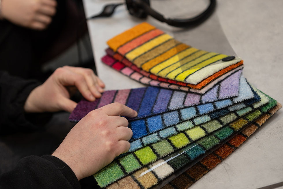 Hands compare stacked colorful carpet samples on a table, with yellow, blue, and green stripes in a showroom-like setting.