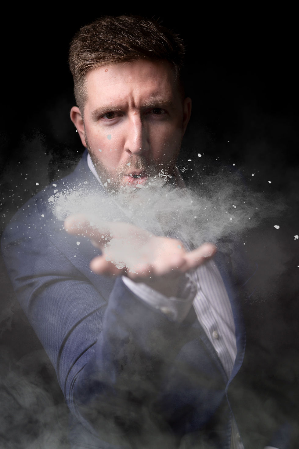 Man in a blue suit blows white powder from his outstretched hand against a black background, with an intense stare.
