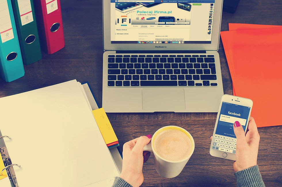 woman's hand holding a coffee and looking at Facebook in her mobile phone whist sitting at her desk with her laptop open and files on her desk