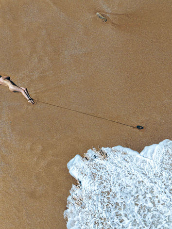 Aerial fine-art nude of a person lying on wet sand near ocean waves in a minimalist coastal composition.