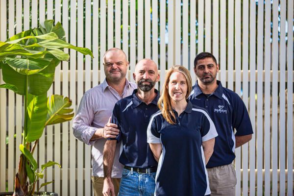 Professional group portrait of four team members standing outdoors in natural light against a white slatted fence, captured by Sydney Commercial Photography.