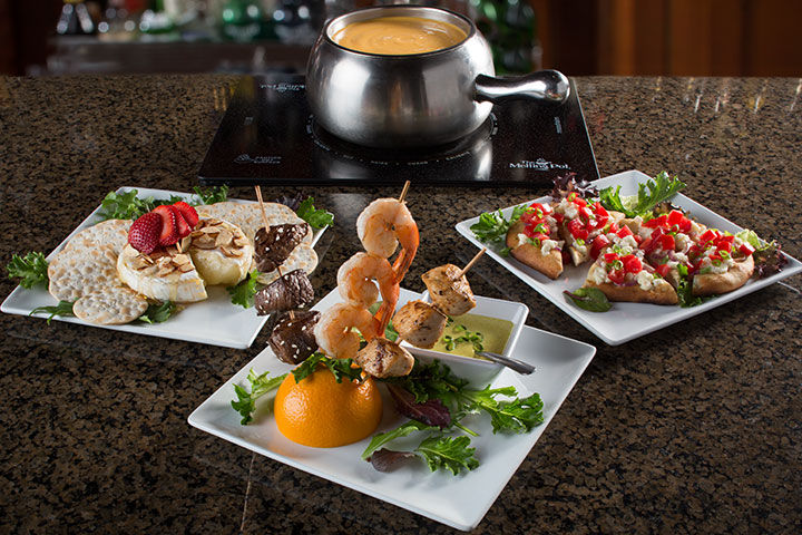 Plates of skewered shrimp, beef, and vegetable dishes on a granite counter with a pot of fondue, surrounded by greens and colorful appetizers.