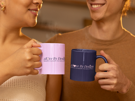 Two people smiling hold pink and blue mugs with "2 Love By Design" logo. Warm indoor setting, cozy and cheerful mood.