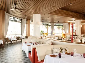 Elegant restaurant interior with curved booths, white tablecloths, and wine glasses. Wood ceiling, patterned tile floor, and large windows. Cozy ambiance.