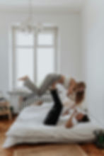 Couple playing on bed, woman balanced on man's feet. Bright room with white walls, wooden floor, and window. Playful and joyful mood.