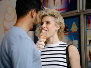 A woman in a striped shirt licks a colorful ice cream cone beside a man, with posters of various ice cream flavors in the background.