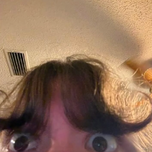 Close-up selfie of curled center-parted bangs swooping upward like a tiny mustache above wide eyes.