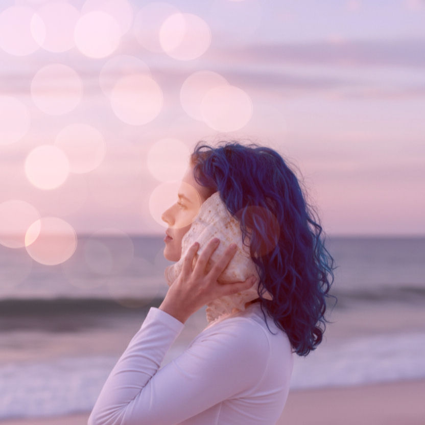 A person with  long hair and soft features holding a large conch shell  to their ear