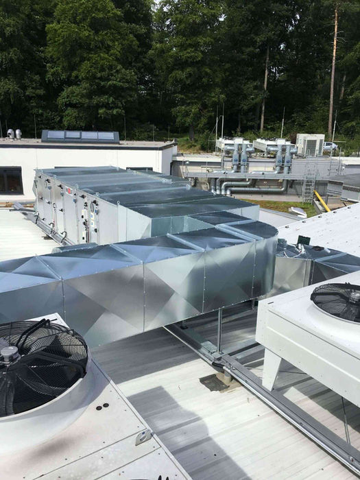 Mehrere weiße Außengeräte, silberne trapezförmige Kanäle und Ventilatoren auf weißem Flachdach mit Bäumen
​