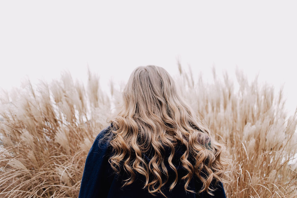A woman with long, curly, blond hair wearing a navy sweater stands with her back to the viewer. She looks at a tall, tan bundle of grasses.