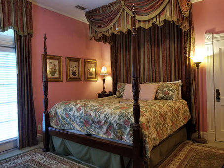 Elegant bedroom with a four-poster bed, floral bedding, and draped canopy. Pink walls, framed art, and warm lamps create a cozy ambiance.