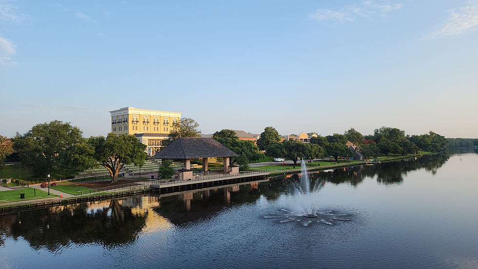 Scene of the beautiful Cane River Lake with a fountain and pavilion. Historic Front Street streetscape is the background. Lush trees and a clear blue sky are reflected in the calm water.