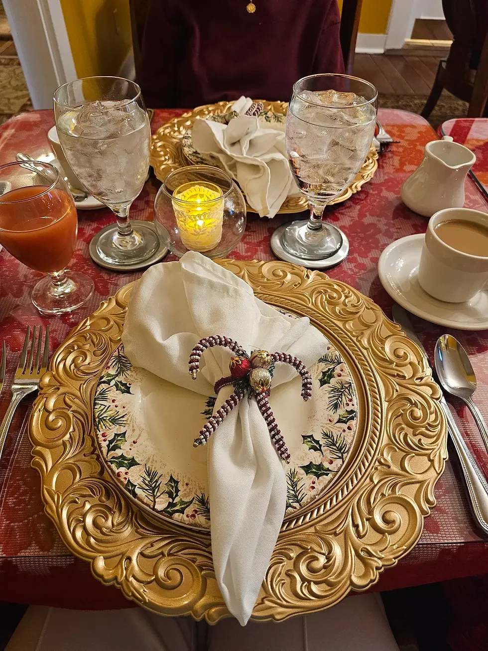 Elegant table setting with gold plates, festive napkin ring, glasses of water, coffee, juice, and a lit candle on a red tablecloth.