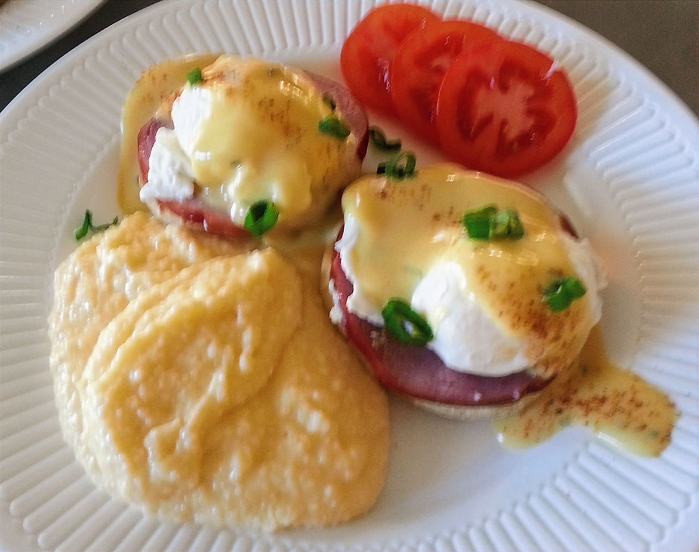 Eggs Benedict with hollandaise and green onions on a white plate, accompanied by grits and sliced tomatoes.