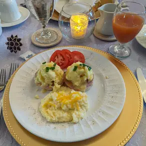 Elegant table setting with plated breakfast of eggs, tomato slices, and grits. Water, juice, and a candle on lavender cloth background.
