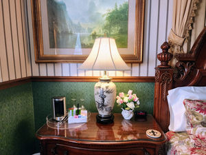 Elegant bedside table with lit lamp, flowers, toiletries tray, and digital clock reading 9:57 beside floral bedding and framed landscape.