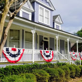 Victorian house with white porch, red, white, and blue bunting, and USA flag. Sign reads "Violet Hill Bed & Breakfast." Tree and bushes in foreground.