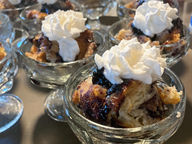A side-angle shot of a gourmet breakfast starter showing warm blueberry croissant bread pudding beautifully presented inside a clear pedestal crystal bowl and crowned with fresh whipped cream.
