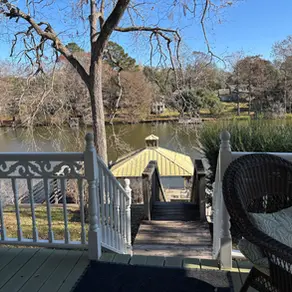 Lake view from porch with white railing. A wicker chair with cushions is in the foreground. Wooden stairs lead to water, surrounded by trees.