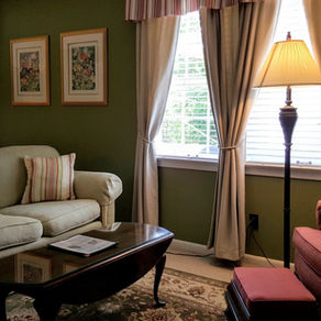 Cozy living room with green walls, beige sofa, red armchair, and lamp. Decorative curtains, art, and coffee table with papers.