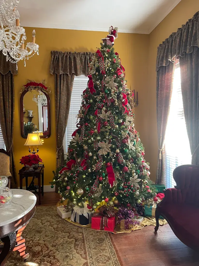 Christmas tree adorned with red ribbons, ornaments, and lights in a yellow room. Presents below, chandelier above, sunlight through curtains.