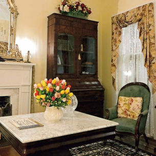 Historic Judge Porter House parlor with an ornate mirror, fireplace, chandelier, antique bookcase, floral chairs, and fresh flowers.