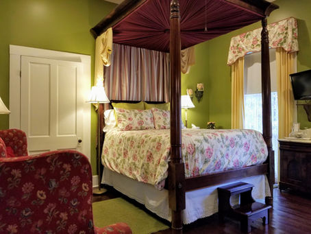Cozy bedroom with a canopy bed, floral bedding, and pink armchair. Green walls, lamps, TV, and window with floral drapes create a warm vibe.