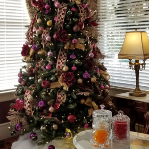 Decorated Christmas tree with red and gold ornaments beside a lit lamp. Elegant drapes frame the window; festive mood.