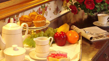 Elegant breakfast setting with tea set, muffins, fruit, and a newspaper on a wooden table. Red roses and floral wallpaper in the background.