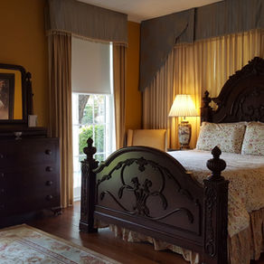 Sumptuous bedroom with ornate dark wood bed, floral bedding, yellow walls, and two lit lamps on nightstands. Draped windows allow soft light in.