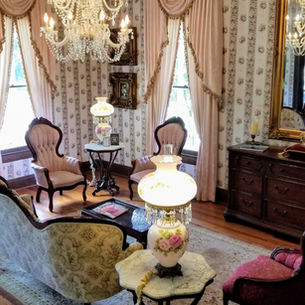 Ornate vintage living room with chandeliers, floral wallpaper, antique furniture, and a large mirror. Warm lighting and elegant decor.