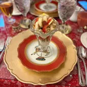 Elegant table setting with a parfait in focus, featuring bananas and strawberries. Red and gold plates on a red lace tablecloth.