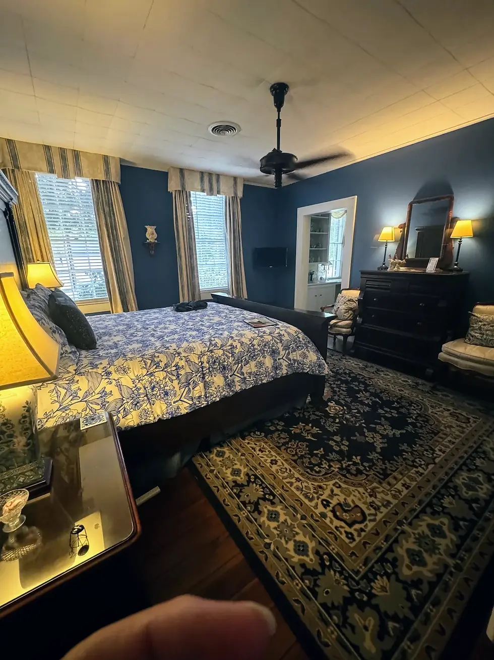 Cozy bedroom with blue walls, floral bedspread, patterned rug, and vintage decor. Lamps glow warmly. Ceiling fan above, window curtains drawn.