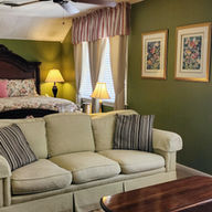 Cozy living room with a beige sofa, red armchair, and wooden coffee table. Green walls, floral rug, striped curtains, and framed art enhance the warmth.