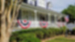 Historic lavender house with American flags and bunting, surrounded by green bushes. Sign reads "Violet Hill Bed & Breakfast." Bright day.