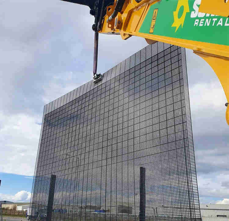 A yellow and green crane lifts a large double 358 mesh fence over an industrial area, utilising the QAB System lifting bracket. 