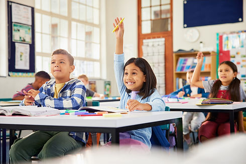 Excited hispanic little girl raising hand in elementary classroom. School children during