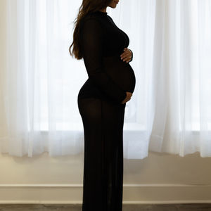 Pregnant woman in black dress, standing by window.