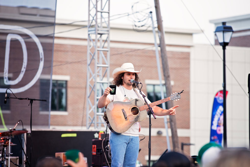 Musician performing outdoors with guitar and cowboy hat.
