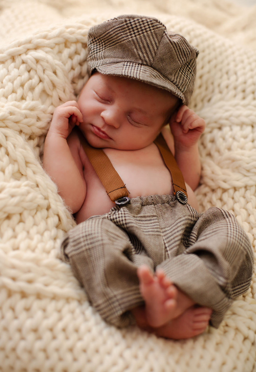 Sleeping baby in suspenders and cap on knitted blanket.