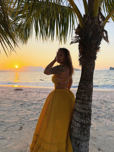 Candice Mary wearing a yellow flowy strapless maxi dress standing on the beach at sunset near a palm tree