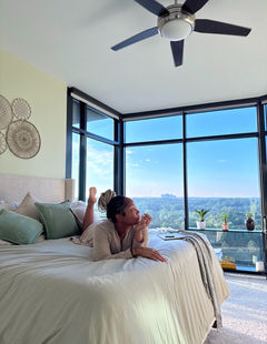 EatMahKandy relaxing on a neatly styled bed in a bright neutral bedroom with large windows, soft bedding, and a cozy modern aesthetic