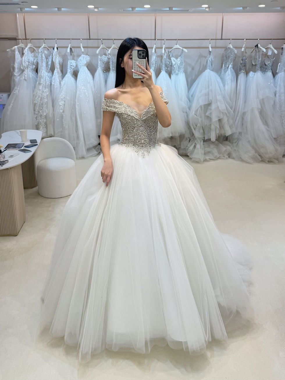 Thumbnail: Off-the-shoulder ball gown with a silver crystal bodice and voluminous white tulle skirt in a bridal boutique.