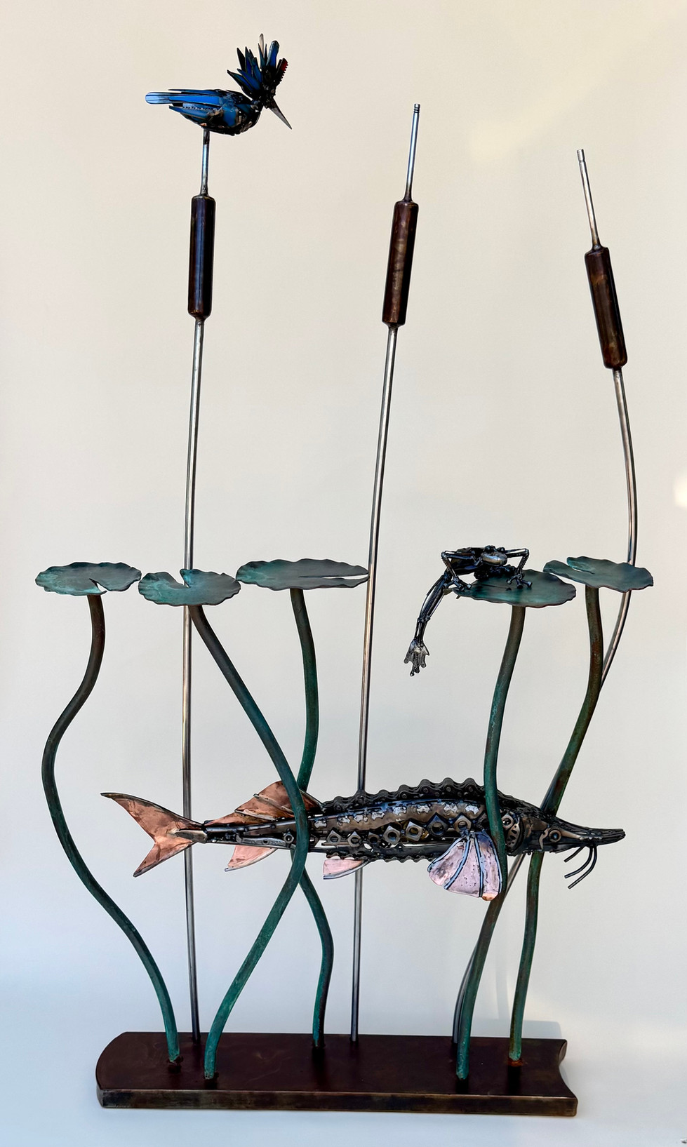 Scrap metal sculptural entry table. A copper and steel sturgeon swims through 5 green lily pad stalks and 3 stainless cat tail stalks.  A frog made from silverware, typewriter parts and salvaged metals climbs onto a lily pad to get away from the sturgeon.  A blue kingfisher, also made from silverware and salvaged metals, perches on one of the cattails.