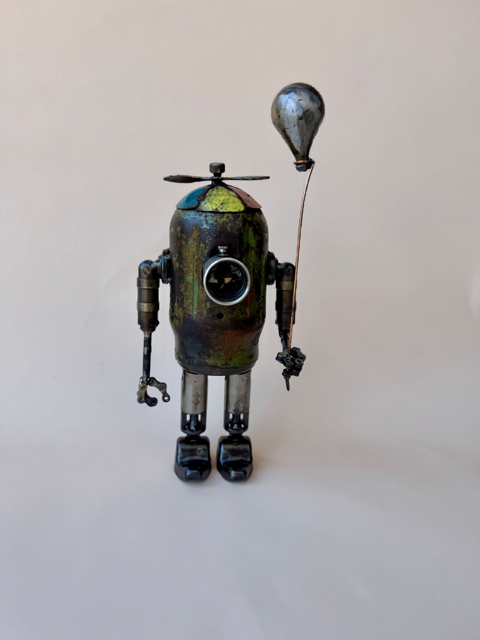 Front view of a small green little boy robot holding a balloon. He has a colorful propeller hat.